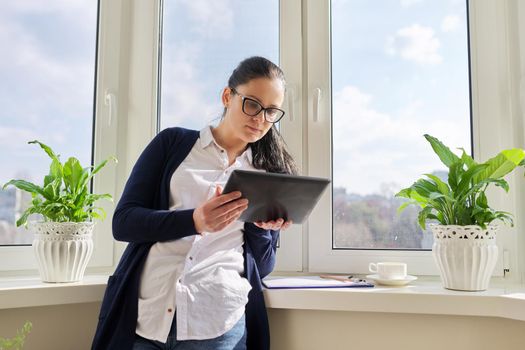 Serious business woman working remotely in her home office, talking on video call, using digital tablet. Freelance, technology, telecommunications, business remotely, video communication near window