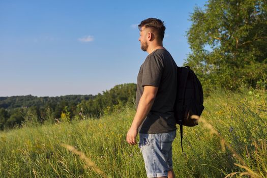 Back view of middle-aged man with backpack looking in distance at natural hills, meadows, sky. Hike, travel, summer, active lifestyle, people 40s age concept