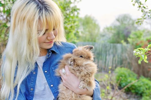 Young blonde woman with decorative rabbit in the spring garden. Female teenager with a pet fluffy bunny. Nature, pets, domestic animals, copy space