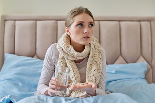 Sick woman with glass of water and pills in her hand, young female sitting at home in bed taking medicine. Medicine, pharmacology, flu season, seasonal diseases, people concept