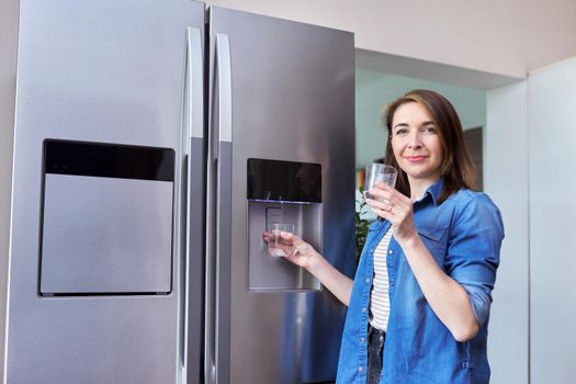 Water dispenser, woman taking cold water into glass from home modern chrome refrigerator in kitchen, dispenser of fridge. Cool water, household, comfort concept