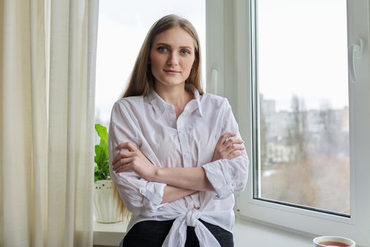 Portrait of young beautiful blonde woman in white shirt near winter autumn window. Smiling 20s blonde female relaxing looking at camera