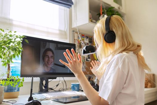 Teen female in headphones talking online with her friend male teenager, using video call on computer, at home at table. Technology, telecommunication, friendship, communication remotely, youth concept