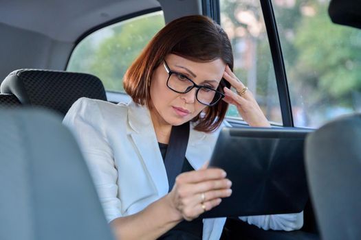 Middle-aged business woman in car in passenger backseat. Serious female in glasses with digital tablet. Business people of mature age, city, emotion fatigue meditation sadness depression frustration