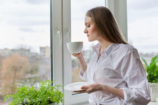 Portrait of young beautiful woman in white shirt with cup of coffee near window. Blonde female relaxing looking out the window of winter autumn city