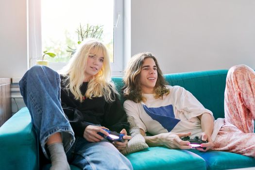 Couple of 16, 17 years old teenage friends playing with video game console, sitting at home on couch, having fun together. Real people, teenagers lifestyle, virtual computer games concept