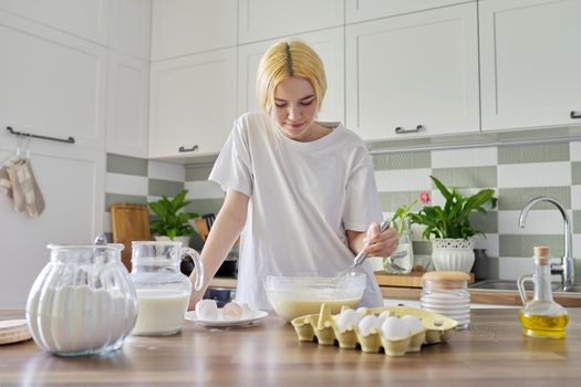 Female teenager preparing food at home, girl in the kitchen, ingredients for making pancakes eggs flour milk sugar on the table. Eating at home, healthy homemade food, teen hobbies