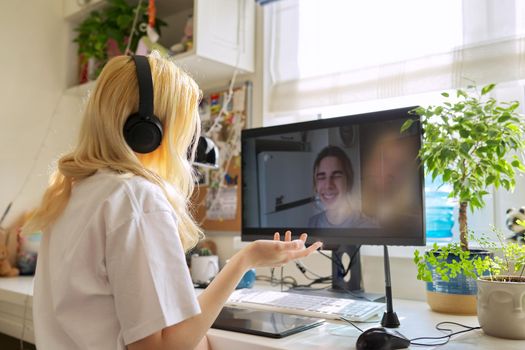 Teen female in headphones talking online with her friend male teenager, using video call on computer, at home at table. Technology, telecommunication, friendship, communication remotely, youth concept