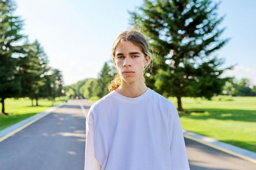 Portrait of handsome teenage guy looking in camera outdoors. Serious hipster teenager with long hair wearing white t-shirt on road on sunny summer day. Youth, adolescence, people, lifestyle concept