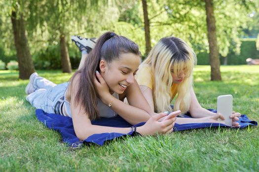Two young female teenagers lying on grass in park with smartphones. Girls looking at phone screen. Youth, adolescents, technology, lifestyle, leisure concept