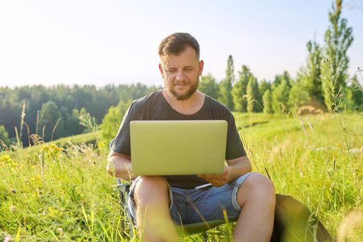 Middle-aged man with laptop in nature. Bearded male sitting on an outdoor chair in meadow in nature, enjoying summer day and using modern technologies for rest and work