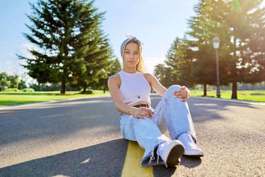 Fashionable trendy teenage hipster female sitting on road in park on sunny summer day. Young stylish model, outdoor. Youth, teens, summer, fashion, street style, lifestyle concept