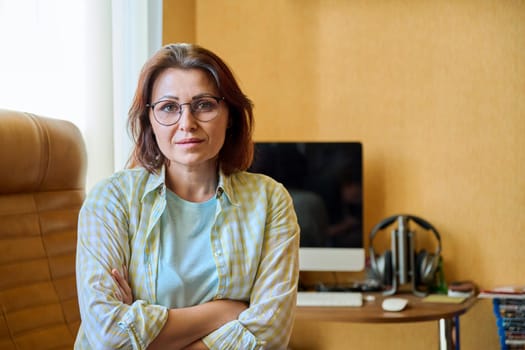 Portrait of confident smiling middle aged woman looking at camera with crossed arms at home workplace, 40s female with glasses sitting on armchair near computer