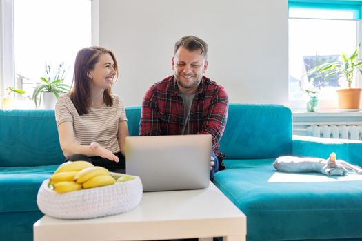 Smiling laughing positive couple 40 years old, husband and wife looking at laptop monitor sitting together at home on sofa in living room, real people life