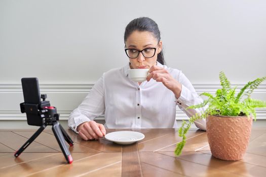 Young woman blogger vlogger recording broadcast at home using smartphone on tripod to communicate with followers. Female at the table with cup of coffee
