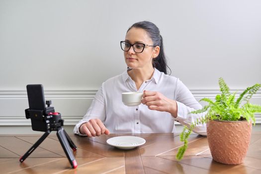 Young woman blogger vlogger recording broadcast at home using smartphone on tripod to communicate with followers. Female at the table with cup of coffee