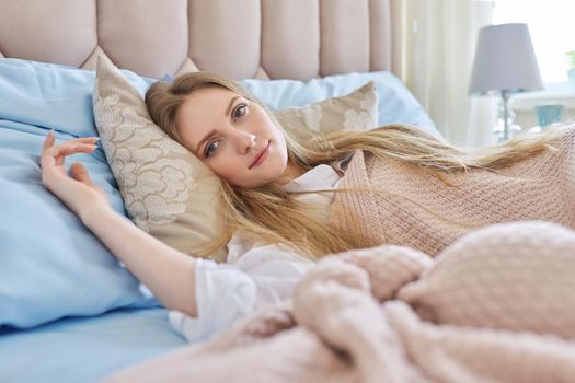 Young beautiful smiling blonde woman resting in bed at home. Close up face looking at camera, blue bed under pink pastel knitted blanket