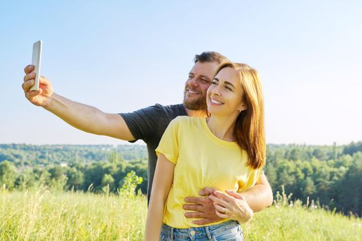 Beautiful happy adult couple taking selfie on smartphone, sunny summer day meadow forest sky background. Relationships, happiness, relaxation, love, people 30s 40s age