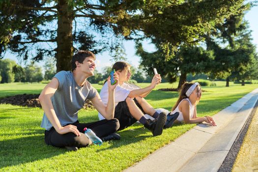 Hipster teenagers having fun in park, sitting on green grass on lawn, recording videos on smartphone, listening to music. Youth, lifestyle, friendship, leisure, technology concept