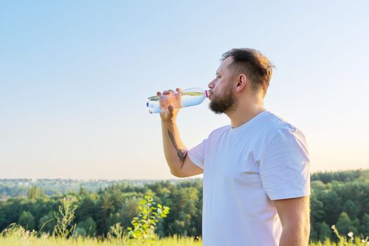 Middle-aged bearded man drinks water from bottle on hot summer day, nature meadow sky hills background. Summer, heat, health concept, copy space