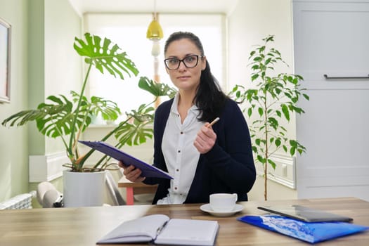 Business woman looking at camera talking, holding papers documents in hands, home interior background. Online consultation, remote work, paperwork, internet, technologies in business, education