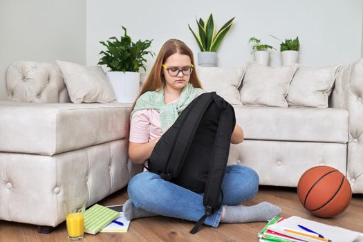 Lifestyle, education children 12, 13 years old concept. Teenage girl sitting at home on the living room floor with backpack, books, school notebooks, basketball