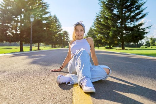 Fashionable trendy teenage hipster female sitting on road in park on sunny summer day. Young stylish model, outdoor. Youth, teens, summer, fashion, street style, lifestyle concept