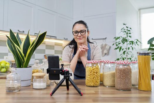 Online broadcast, blog about food, woman in an apron with jars of cereals noodles flour, looking at smartphone camera, telling and showing about food storage, organizing kitchen