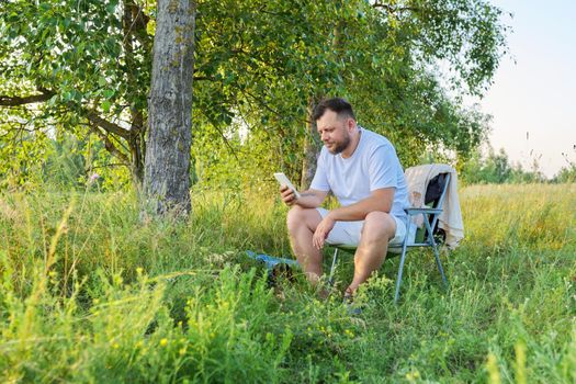 Middle-aged bearded man having a rest sitting on an outdoor chair in nature, using a smartphone, summer sunny day background