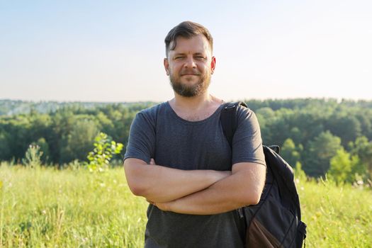 Portrait of confident smiling man in his 40s with folded arms looking in camera. Bearded male with backpack on sunny summer day, sky meadow nature background