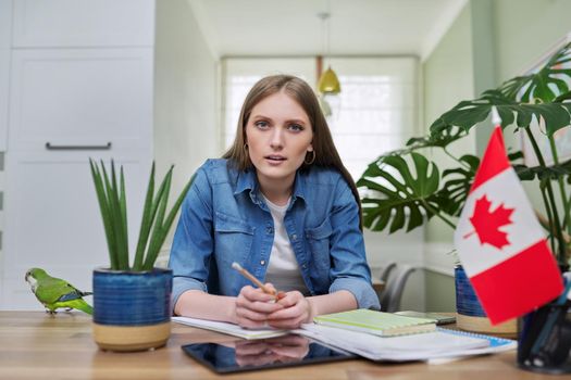 Canada, education, study, students concept. Online training, distance lessons, e-learning, female teenager university student sitting at home looking at webcam, close-up face, Canadian flag on table