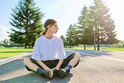 Portrait of handsome teenage guy, profile view outdoors, copy space. Serious hipster teenager with long hair wearing white t-shirt on road on sunny summer day. Youth, adolescence, people, lifestyle concept