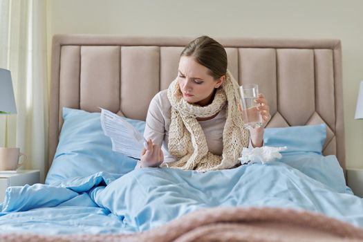 Young sick woman at home in bed, female with glass of water reading instructions and recommendations for medication. Season of colds, viral diseases, health, medicine, pharmacology, people concept.