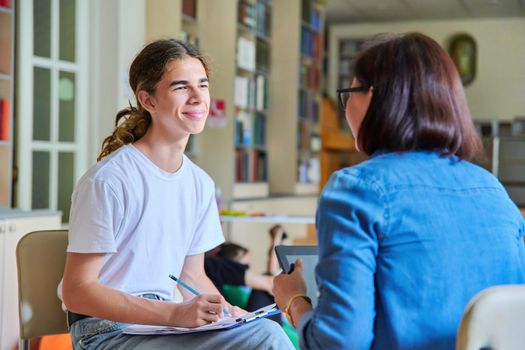 Woman school psychologist teacher talking and helping student, male teenager, inside the library. Mental health of adolescents, psychology, social issues, professional help of counselor