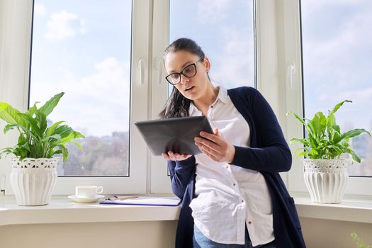 Serious business woman working remotely in her home office, talking on video call, using digital tablet. Freelance, technology, telecommunications, business remotely, video communication near window