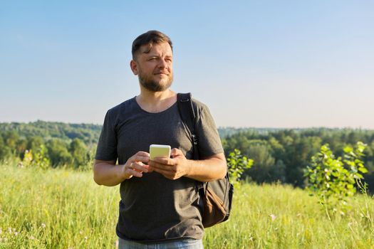 Middle-aged bearded man with backpack using smartphone. Summer sunny day, nature, sky background. Hike, adventure, vacation, technology, communication, leisure people concept