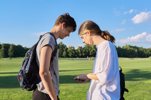 Teenagers friends talking looking at smartphone screen. Two guys 16, 17 years old with smartphone outdoors, on road on sunny summer day. Technology, lifestyle, friendship, youth, adolescence concept