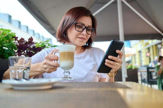 Smiling middle aged woman sitting in an outdoor cafe with cup of coffee and looking at smartphone screen. Coffee break, people 40s age, urban style, leisure, lifestyle, technology concept