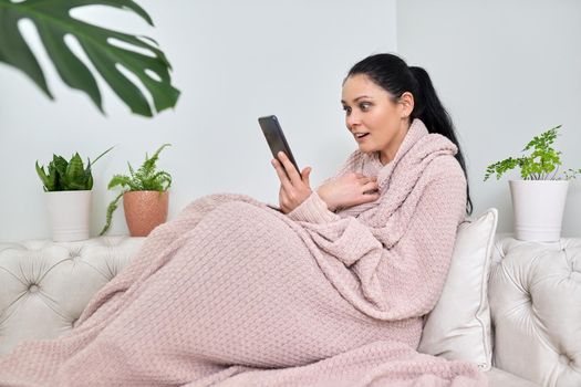 Surprised emotional middle-aged woman using smartphone for video call, female in autumn winter season under warm knitted blanket sitting on sofa in living room. Video, online, virtual meetings, communication