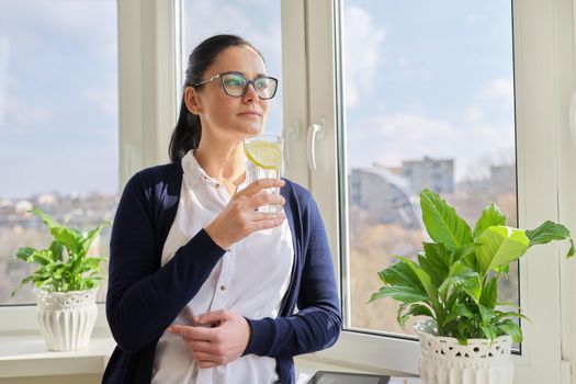 Business woman with glass of water with lemon. Middle-aged female in glasses, white shirt cardigan near window, resting enjoying healthy vitamin water