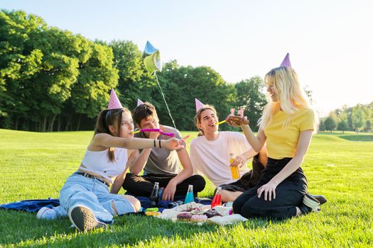 Birthday party. Teenager girl with cake with candles 17, celebrating birthday with friends, teenagers in festive hats sitting on grass in park, summer sunny day