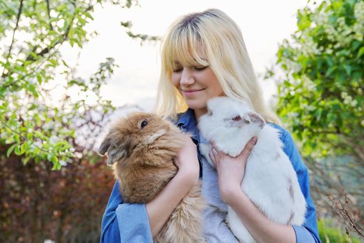 Beautiful smiling girl teenager with a couple of decorative rabbits in their hands. Blonde female in denim clothes with two bunnies in a spring blooming garden