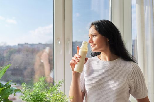 Woman drinking milk drink from bottle standing near window, milk yogurt dairy healthy drinks, copy space. Healthy food, diet, natural products