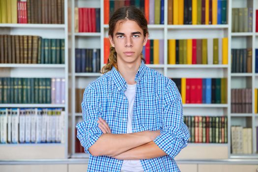 Single portrait of serious confident male student teenager with crossed arms looking at camera in the library. School, education, knowledge, adolescence concept
