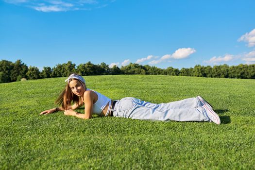 Girl hipster teenager lying on grass, green lawn and blue sky background. Summer, vacation, nature, beauty, relax, lifestyle, leisure, youth concept