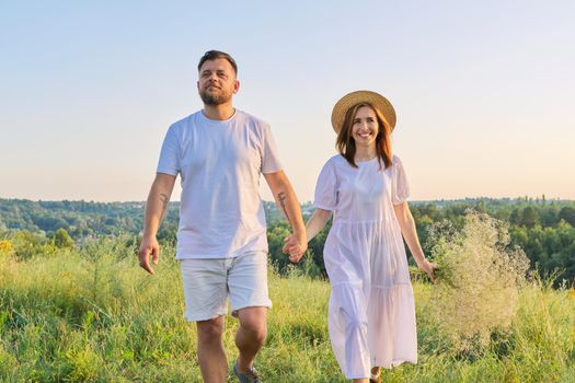 Happy romantic middle age couple, smiling man and woman walking together holding hands, female with bouquet in white dress hat, natural landscape in sunset light background. Relationships, love, dating, wedding anniversary, people 30s 40s age