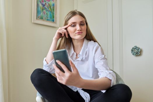 Emotional laughing young beautiful woman looking at smartphone screen, relaxing sitting at home in chair. Video call, emotional communication, joy, happiness, surprise, technology in everyday life