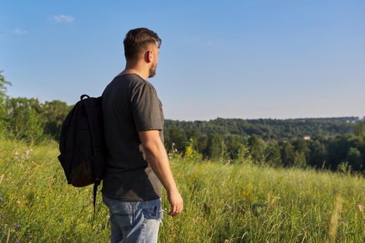 Back view of middle-aged man with backpack looking in distance at natural hills, meadows, sky. Hike, travel, summer, active lifestyle, people 40s age concept
