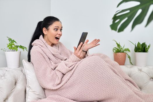 Surprised emotional middle-aged woman using smartphone for video call, female in autumn winter season under warm knitted blanket sitting on sofa in living room. Video, online, virtual meetings, communication