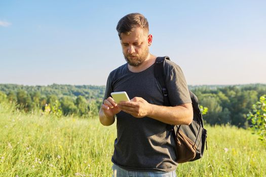 Middle-aged bearded man with backpack using smartphone. Summer sunny day, nature, sky background. Hike, adventure, vacation, technology, communication, leisure people concept